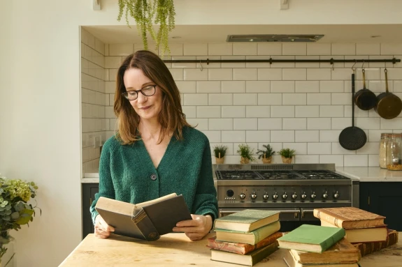 Susie Dent sitting at kitchen table reading a book.